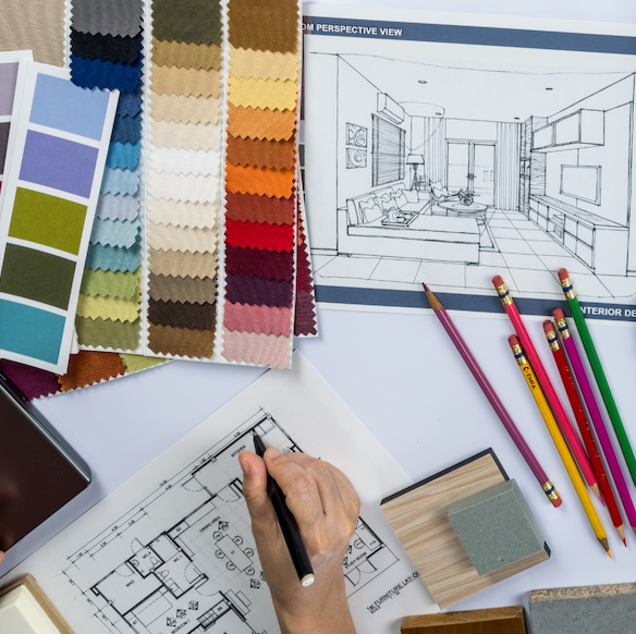 Interior designer’s desk with fabric swatches, color charts, plans and a perspective sketch; pencils and sketchbook visible.
